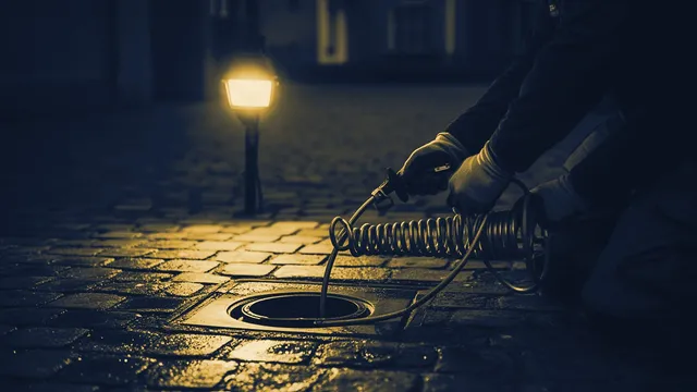 Gehandschoende handen voeren een ontstoppingsveer in een afvoerputje, avondlicht